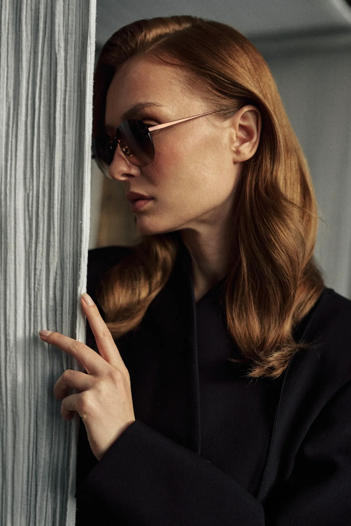 Side profile of a woman in a black coat by a pale curtain, wearing KAIROS Amber brown titanium sunglasses with brown gradient lenses