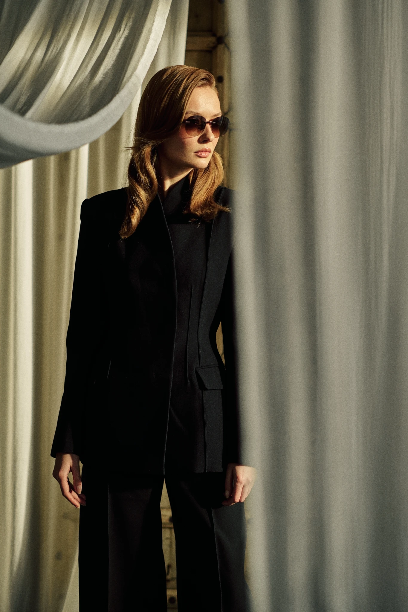 Woman in a black tailored suit between pale curtains, wearing KAIROS Amber sunglasses with brown gradient lenses